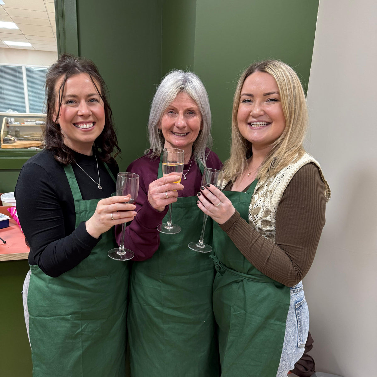 Three women standing together indoors, each wearing a green apron and holding a champagne flute. The setting appears to be a kitchen or workshop with green walls and wood flooring.