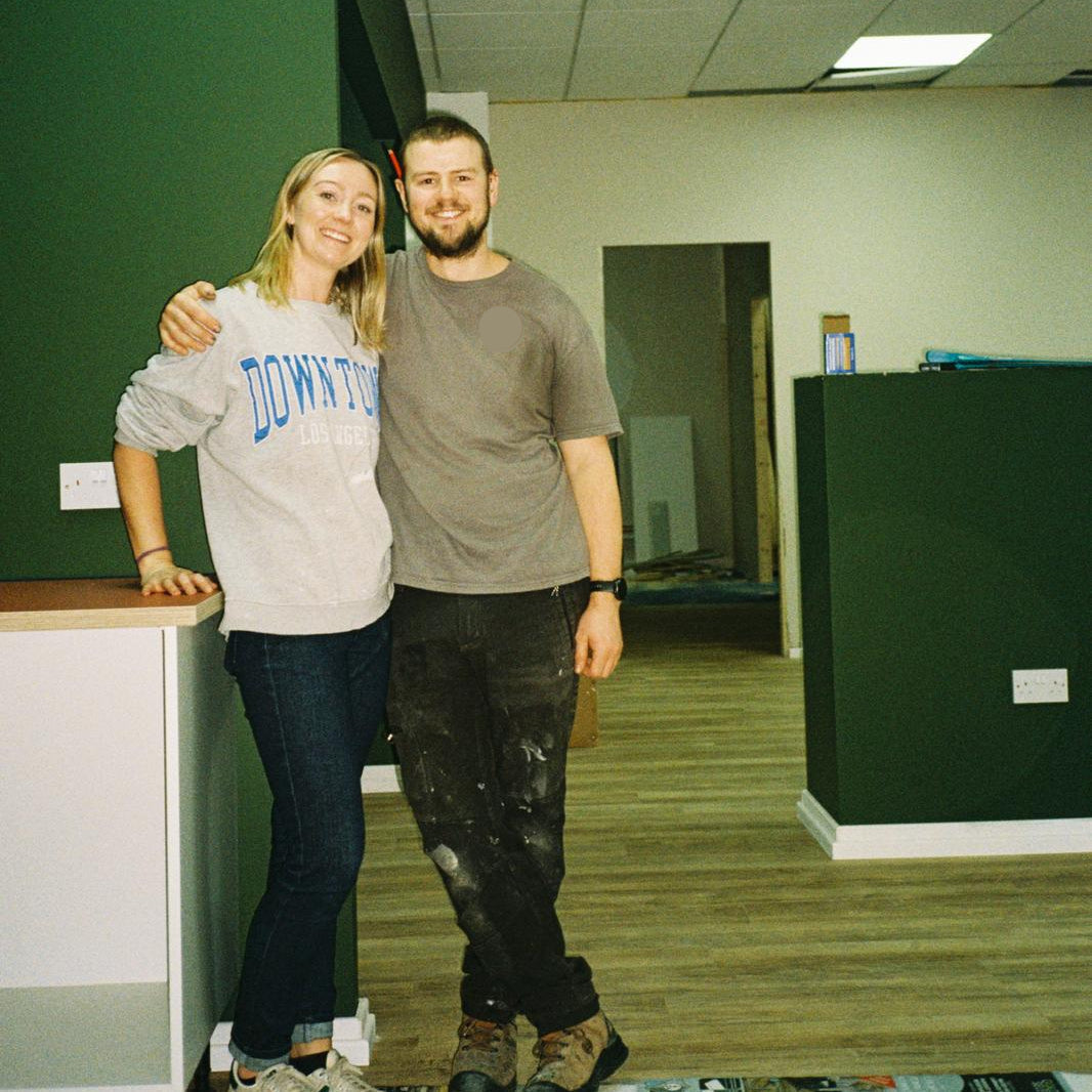 A man and a woman standing together in a room with dark green walls, wood flooring, and a drop ceiling. The woman wears a light gray sweatshirt with 'DOWNTOWN' printed in blue, dark jeans, and sneakers.