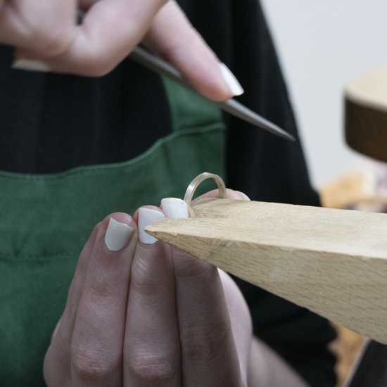 A close-up of a person's hands holding a ring on a wooden ring stand, with a metal file or tool in the air above. The person is wearing a dark green apron over a black top, and a workshop table is visible in the softly blurred background.