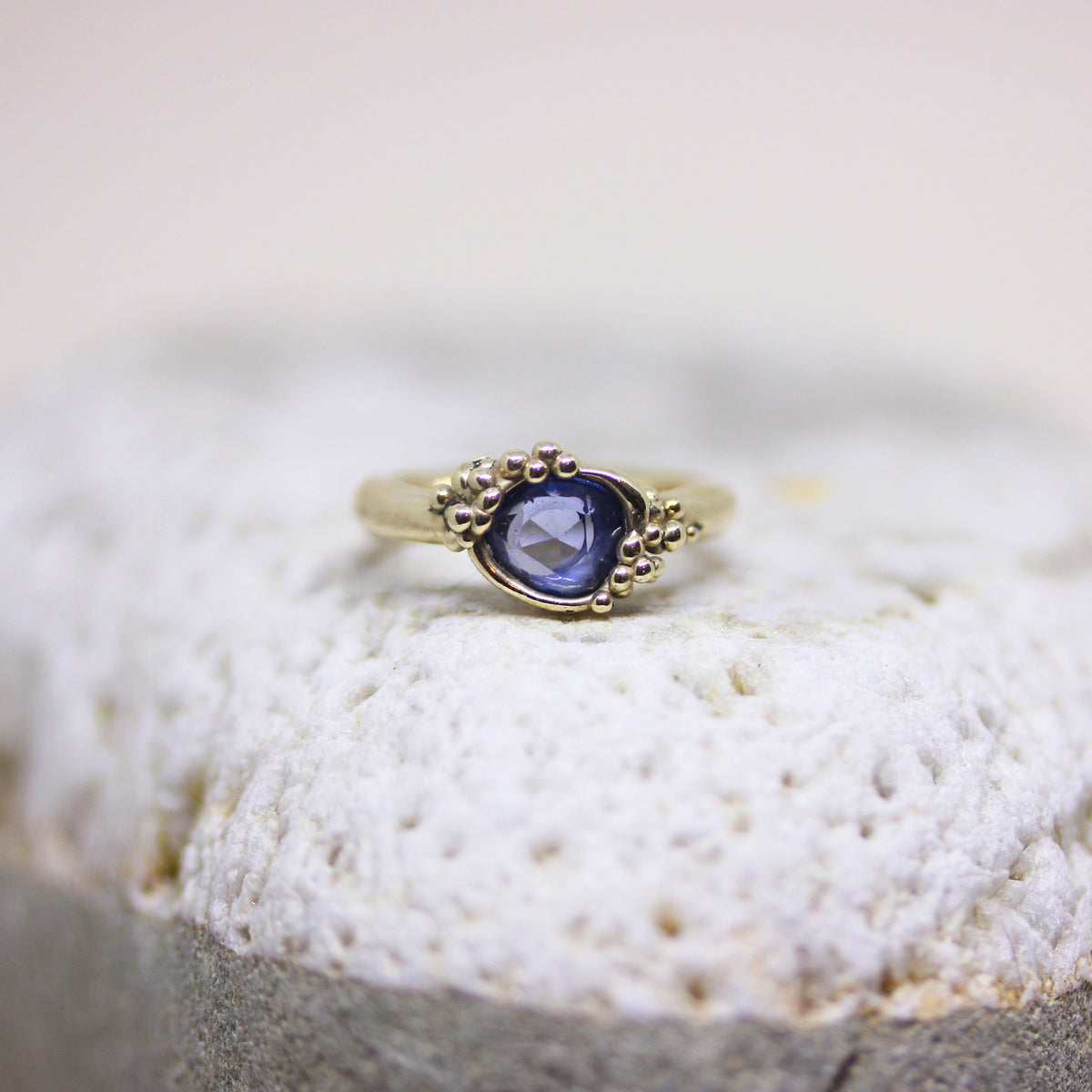 A gold ring with a round blue gemstone and decorative bead accents on the shoulders, resting on a textured white stone surface.