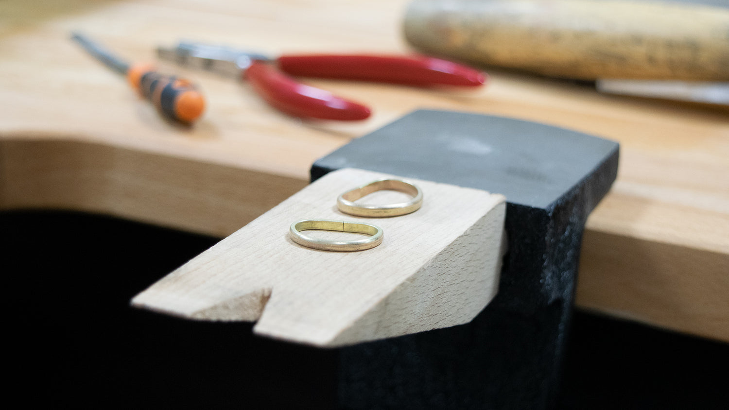 Two plain gold wedding bands displayed on a small wooden block in the foreground, with a black anvil and blurred workshop tools in the background.