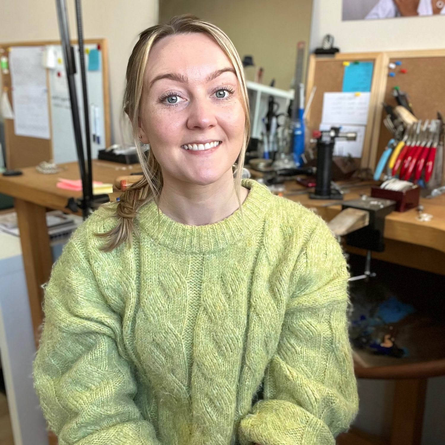 A smiling blonde woman wearing a chunky light green cable-knit sweater, seated in a workshop or craft studio with workbenches and tools in the background.