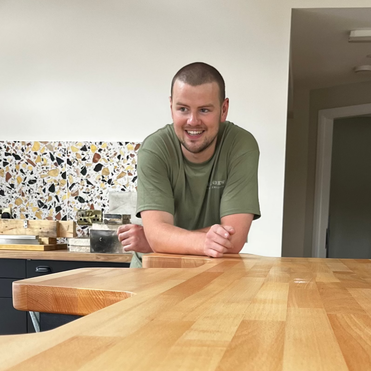A man in an olive green t-shirt leans on a large, polished wooden kitchen island in a modern kitchen setting with a terrazzo backsplash.