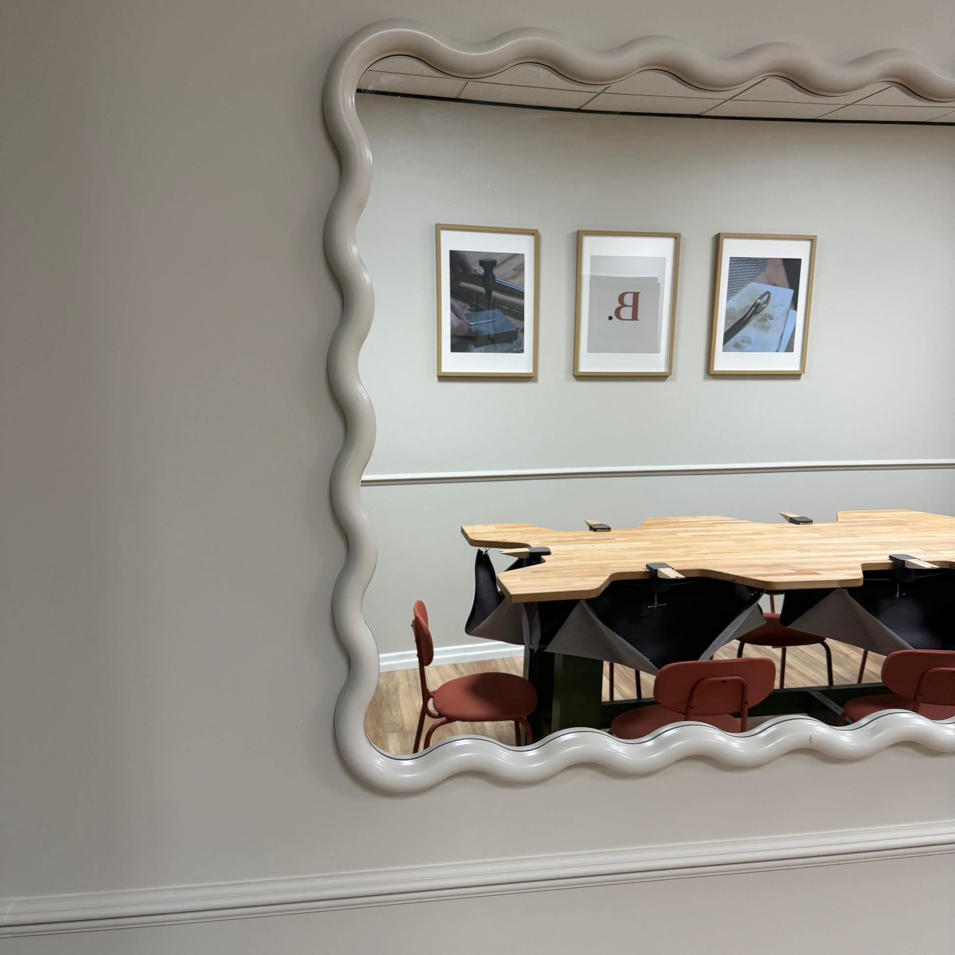 A large wavy-edged mirror mounted on a light gray wall, reflecting a jewellery studio with a wooden jewellers bench, chairs, and three framed pictures on the wall.