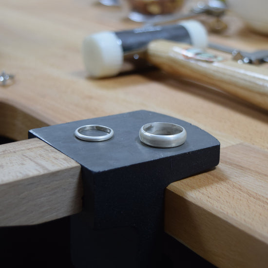 A close-up of a black metal ring clamp mounted on a wooden workbench, holding two silver wedding bands. A wooden-handled tool and other workshop items are blurred in the background.