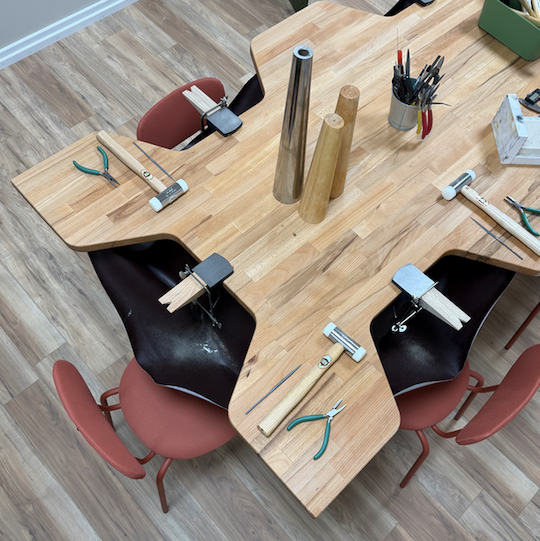 An overhead view of a modern, geometric wooden workbench with integrated tool holders, surrounded by black and terracotta chairs on a wood-look floor. Various woodworking tools are laid out on the table.