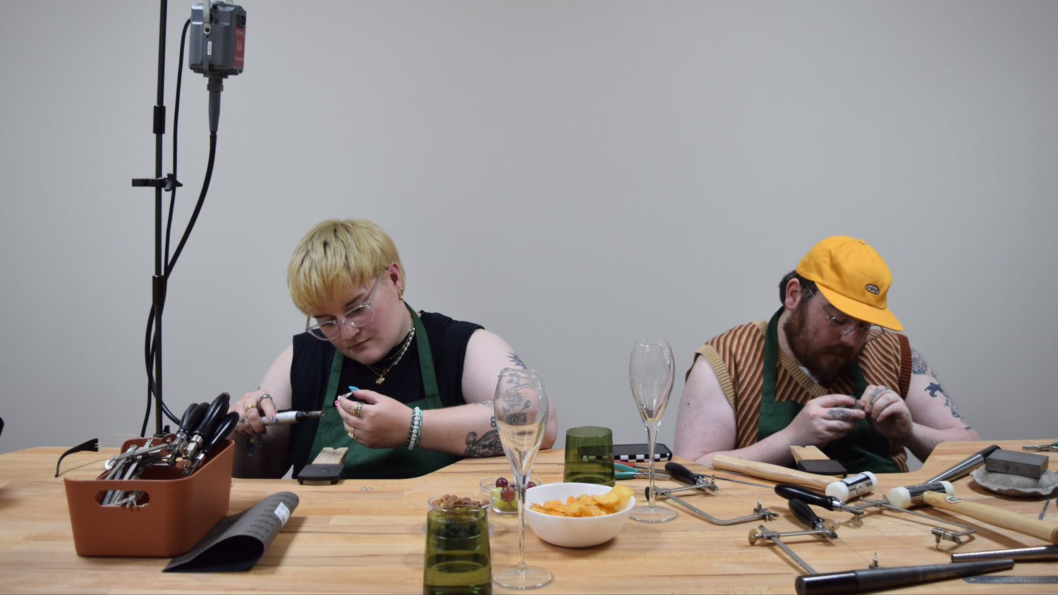 Two people in matching green aprons work at a wooden table filled with tools, glasses, and small objects, under a studio light stand against a plain gray wall.